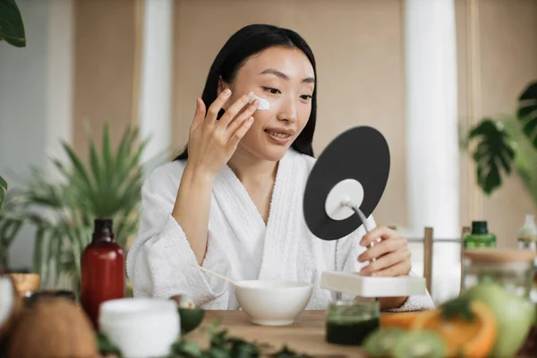 身穿白色浴衣的美丽健康的亚洲女人坐在木制桌旁 在家里用各种配料准备天然化妆品 在碗中搅拌 同时准备奶油并涂在脸上 — 图库照片