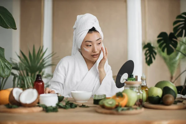 身穿白色浴衣和毛巾的健康的亚洲女人坐在木制桌旁 在家里用各种配料准备天然化妆品 在碗中搅拌 同时准备奶油并涂在脸上 — 图库照片