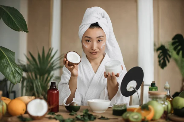 年轻的亚洲女人拿着一半椰子 为健康的皮肤自制奶油 许多自制化妆品的配料躺在木制桌子上 — 图库照片