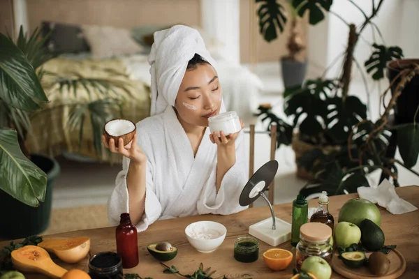 身穿白色浴衣和毛巾的年轻亚洲女人 拿着一半椰子 为健康的皮肤自制奶油 许多自制化妆品的配料躺在木制桌子上 — 图库照片