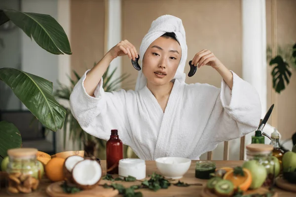 头戴毛巾 身穿浴衣 面带微笑的亚洲女人坐在桌旁 手里拿着胶原蛋白片 年轻女性在异国情调的工作室里用自制化妆品进行日常美容 — 图库照片