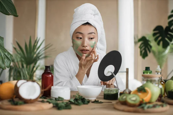 迷人的亚洲女人看着坐在桌边的镜子 桌上有自制化妆品配料 脸上涂着绿色保湿面膜 做抗衰老手术的年轻女士 — 图库照片