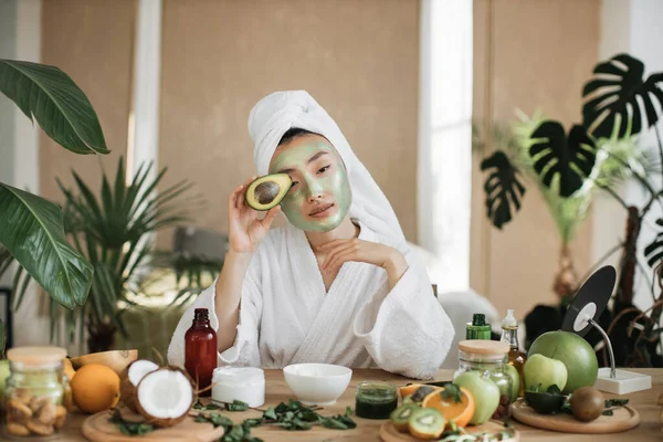 迷人的亚洲女人坐在餐桌旁 在脸上涂上绿色保湿面膜 配上自制化妆品 手握新鲜鳄梨 — 图库照片