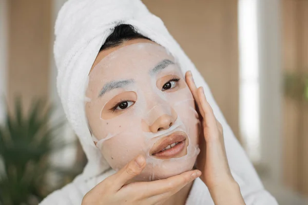Healthy young asian female with cotton mask sheet on face and towel on head over exotic background. Pretty woman doing skin care procedures indoors at home.