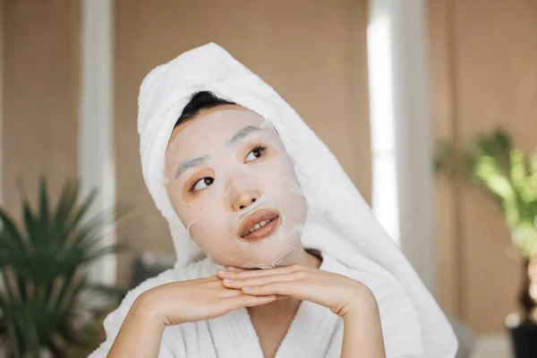 在异国情调的家庭工作室里做皮肤护理手术的漂亮女人 健康的年轻亚洲女人 脸上戴着棉制口罩 头上披着毛巾 背景异国情调 — 图库照片