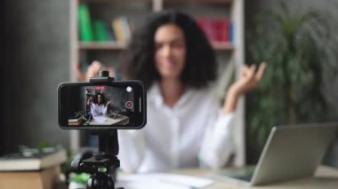 Bulanık, rahat ve rahat genç bir kadın iş yerinde modern bir dizüstü bilgisayarla oturuyor ve video blogu kaydederken gözleri kapalı meditasyon yapıyor. Tripoda sabitlenmiş modern akıllı telefona odaklan.