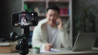 Akıllı telefon ekranına odaklan, akıllı telefondan konuşan ve video kaydına video kaydederken günlük giyinen çekici Asyalı adam. Erkek blogcu evde oturuyor ve canlı yayın yapıyor.