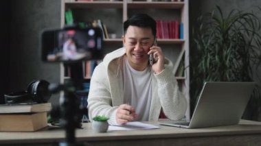 Sıradan giyinen çekici Asyalı adam akıllı telefondan konuşuyor, video bloğu için video kaydederken rapor yazıyor. Erkek blogcu evde oturuyor ve canlı yayın yapıyor.
