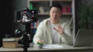 Ekrana odaklan, modern telefonda video kaydederken günlük kıyafetler içinde konuşan ve el kol hareketi yapan yakışıklı Asyalı adam. Erkek blogcu evde oturuyor ve canlı yayın yapıyor.