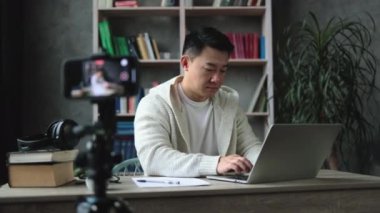 Çekici Asyalı erkek, gündelik giysiler içinde dizüstü bilgisayarda çalışıyor ve dijital akıllı telefondan video kaydediyor. Serbest ve modern teknoloji kavramı.