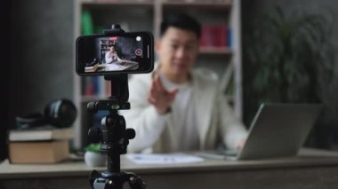 Akıllı telefon ekranına odaklan. Akıllı telefonuyla video kaydederken laptopta rapor yazan hoş bir genç adam. Asyalı erkek serbest yazar evde kalırken uzaktan çalışıyor..