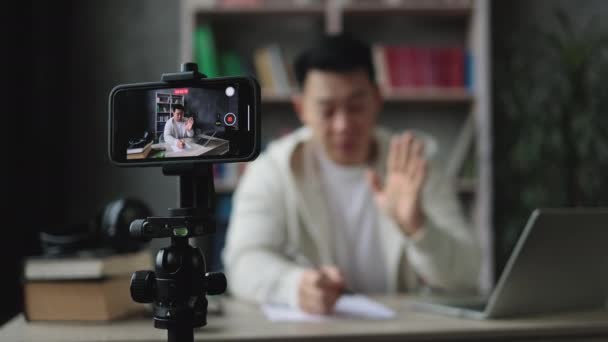 专注于智能手机屏幕 微笑的亚洲男人在现代智能手机相机前说话和做手势 坐在现代家居的背景下写报告 直播概念 — 图库视频影像