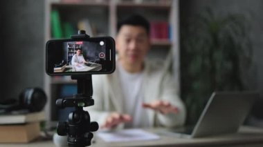 Sıradan giysiler içinde gülümseyen Asyalı adam kitap, not ve dizüstü bilgisayarla oturmuş video blogu kaydediyor ve başparmağını gösteriyor. Uzaktan öğrenim sırasında evde çalışan kadın öğretmen.