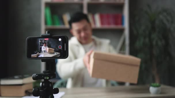 专注于智能手机屏幕快乐的亚裔男子在休闲穿着录象带相机 同时打开礼品盒 男性博主与社交网络订户分享他的情绪 — 图库视频影像