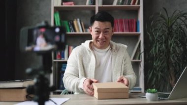 Mutlu Asyalı adam yeni akıllı telefonuyla paket kutusunu açarken modern telefon kamerasıyla video çekiyor. İnsan kavramı, teknoloji ve blog yazma.