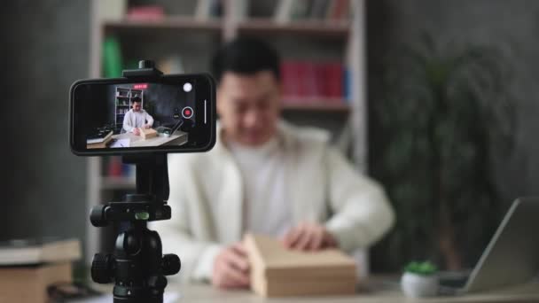 スマートフォンの画面に焦点を当て 新しいスマートフォンで小包ボックスを開きながら 現代の電話カメラでビデオを撮影快適なアジアの男 ブログの概念 — ストック動画