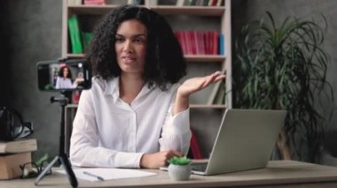 Beyaz gömlekli çok uluslu bir kadın tripodla modern telefondan video kaydederken konuşuyor ve el kol hareketi yapıyor. Kadın blogcu evde oturuyor ve canlı yayın yapıyor.