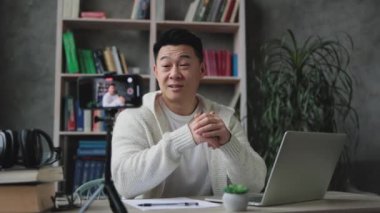 Modern telefondan tripodla video kaydederken günlük kıyafetlerle konuşan ve el kol hareketi yapan çekici Asyalı bir adam. Erkek blogcu evde oturuyor ve canlı yayın yapıyor.