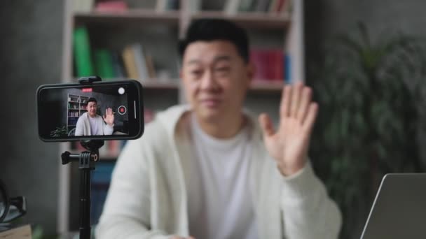 专注于智能手机屏幕 穿着休闲装的亚洲人在三脚架上记录现代手机视频时说话和做手势 男性博主坐在家里做现场直播 — 图库视频影像