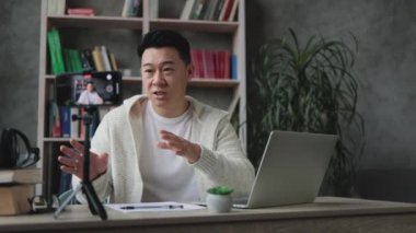 Modern telefondan tripodla video kaydederken günlük kıyafetlerle konuşan ve el kol hareketi yapan çekici Asyalı bir adam. Erkek blogcu evde oturuyor ve canlı yayın yapıyor.