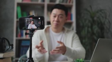 Akıllı telefon ekranına odaklan, sıradan giyinmiş Asyalı adam konuşurken ve el kol hareketi yaparken modern telefondan tripodla video kaydet. Erkek blogcu evde oturuyor ve canlı yayın yapıyor.