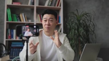 Gündelik kıyafetlerle gülümseyen Asyalı adam kitap, not ve dizüstü bilgisayarla oturmuş video blogu kaydediyor ve imzasını gösteriyor. Uzaktan öğrenim sırasında evde çalışan erkek öğretmen..