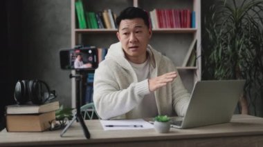 Akıllı telefonuyla tripodla video kaydederken laptopta çalışan hoş Asyalı genç bir adam. Yakışıklı bir erkek, evde otururken uzaktan çalışıyor..