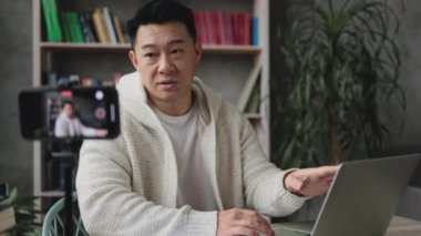Akıllı telefonuyla tripodla video kaydederken laptopta çalışan hoş Asyalı genç bir adam. Yakışıklı bir erkek, evde otururken uzaktan çalışıyor..
