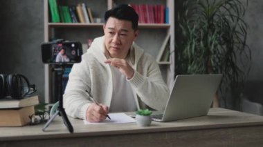 Gülümseyen Asyalı adam tripodla modern akıllı telefon kamerasının önünde konuşuyor ve el kol hareketi yapıyor, modern evin arka planında otururken rapor yazıyor. Canlı yayın kavramı.