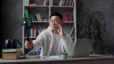 Modern telefondan video kaydederken günlük kıyafetlerle konuşan ve el kol hareketi yapan yakışıklı Asyalı genç adam. Erkek blogcu evde oturuyor ve canlı yayın yapıyor.