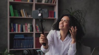 Beyaz tişörtlü, akıllı telefonunda çubuk kullanarak selfie çeken çekici, melez genç bir kadının yakın portresi. Kadın blogcu evde oturuyor ve canlı yayın yapıyor.