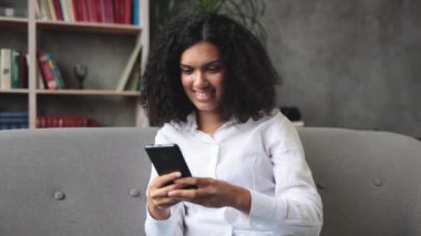 Gündelik giysiler içinde çok uluslu güzel bir kadın modern akıllı telefonuyla rahat bir koltukta oturuyor. Evde boş zamanlarında internette sörf yapan gülümseyen bir kadın..