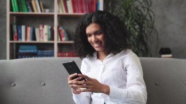 Gündelik giysiler içinde çok uluslu güzel bir kadın modern akıllı telefonuyla rahat bir koltukta oturuyor. Evde boş zamanlarında internette sörf yapan gülümseyen bir kadın..