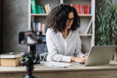 Beyaz tişörtlü, çok uluslu güzel bir bayan dizüstü bilgisayarda çalışıyor ve dijital akıllı telefondan video kaydediyor. Serbest ve modern teknoloji kavramı.