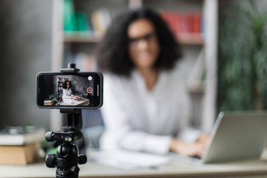 Akıllı telefonlara odaklan, video kaydedilirken laptopta rapor yazan hoş bir genç kadın. Evde otururken uzaktan çalışan çok uluslu kadın serbest çalışan..