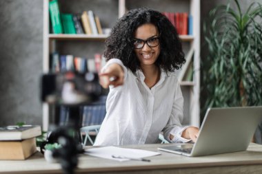 Beyaz gömlekli çok uluslu bir kadın modern telefonda video kaydederken konuşuyor ve el kol hareketi yapıyor. Kadın blogcu evde oturuyor ve canlı yayın yapıyor.