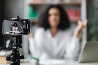 Bulanık, rahat ve rahat genç bir kadın iş yerinde modern bir dizüstü bilgisayarla oturuyor ve video blogu kaydederken gözleri kapalı meditasyon yapıyor. Tripoda sabitlenmiş modern akıllı telefona odaklan.