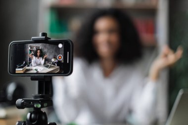 Bulanık, rahat ve rahat genç bir kadın iş yerinde modern bir dizüstü bilgisayarla oturuyor ve video blogu kaydederken gözleri kapalı meditasyon yapıyor. Tripoda sabitlenmiş modern akıllı telefona odaklan.