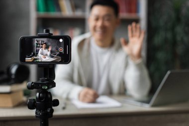Ekrana odaklan, modern telefonda video kaydederken günlük kıyafetler içinde konuşan ve el kol hareketi yapan yakışıklı Asyalı adam. Erkek blogcu evde oturuyor ve canlı yayın yapıyor.
