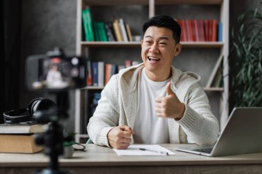 Sıradan giysiler içinde gülümseyen Asyalı adam kitap, not ve dizüstü bilgisayarla oturmuş video blogu kaydediyor ve başparmağını gösteriyor. Uzaktan öğrenim sırasında evde çalışan erkek öğretmen..