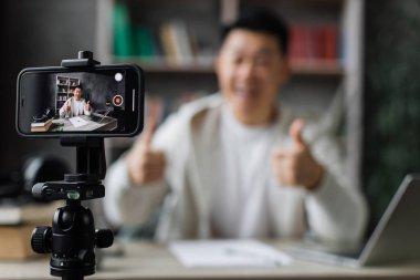 Sıradan giysiler içinde gülümseyen Asyalı adam kitap, not ve dizüstü bilgisayarla oturmuş video blogu kaydediyor ve başparmağını gösteriyor. Uzaktan öğrenim sırasında evde çalışan erkek öğretmen..