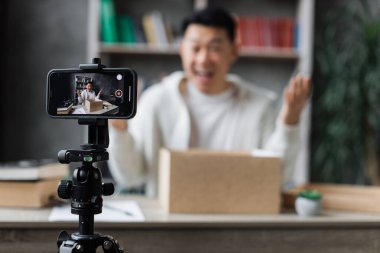 Akıllı telefon ekranına odaklan, mutlu Asyalı erkek blogcu şirketlerin hediye kutularını açıyor ve canlı yayın yapıyor. İçerik yaratmak için akıllı telefon kamerası kullanan çekici bir adam..