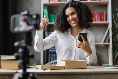 Modern telefon kamerasıyla video çeken çok uluslu bir kadın yeni akıllı telefonuyla paket kutusunu açarken baş parmağını gösteriyor. İnsan kavramı, teknoloji ve blog yazma.