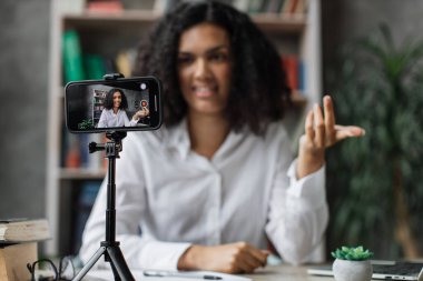 Beyaz gömlekli çok uluslu bir kadın tripodla modern telefondan video kaydederken konuşuyor ve el kol hareketi yapıyor. Kadın blogcu evde oturuyor ve canlı yayın yapıyor.