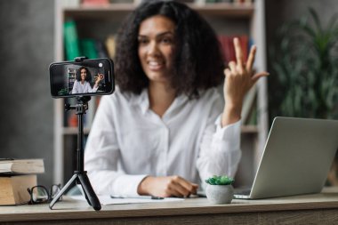 Ekrana odaklan, beyaz gömlekli çekici kadın tripodla modern telefondan video kaydederken konuşuyor ve el kol hareketi yapıyor. Kadın blogcu evde oturuyor ve canlı yayın yapıyor.