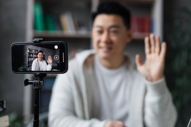 Ekrana odaklan, gündelik giysiler içindeki çekici Asyalı adam tripodla modern telefondan video kaydederken konuşuyor ve el kol hareketi yapıyor. Erkek blogcu evde oturuyor ve canlı yayın yapıyor.