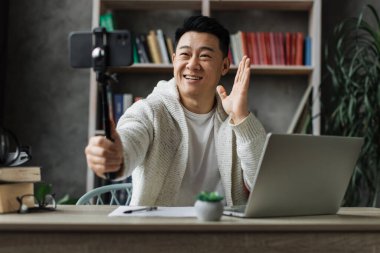 Modern telefondan video kaydederken günlük kıyafetlerle konuşan ve el kol hareketi yapan yakışıklı Asyalı genç adam. Erkek blogcu evde oturuyor ve canlı yayın yapıyor.