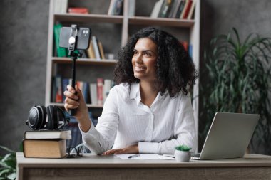 Beyaz tişörtlü çok uluslu çekici bir kadın modern telefondan selfie çekiyor. Kadın blog yazarı evde masada oturuyor ve canlı yayın yapıyor..