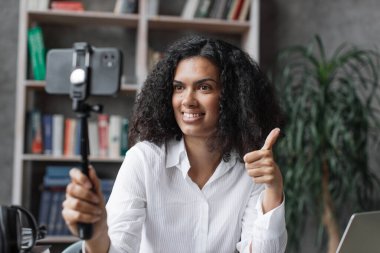 Beyaz tişörtlü çok uluslu bir kadın modern telefondan video kaydederken konuşuyor ve el kol hareketi yapıyor. Kadın blogcu evde oturuyor ve canlı yayın yapıyor..