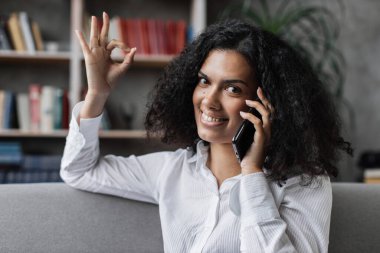 Rahat bir kanepede dinlenirken akıllı telefondan konuşan gündelik giyimli neşeli genç bayan. Evde boş zamanlarında cep telefonu kullanan genç bir kadın..
